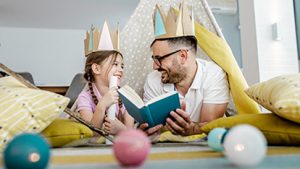 Father and his little girl are on the floor surrounded by toys, he is reading a story to his girl
